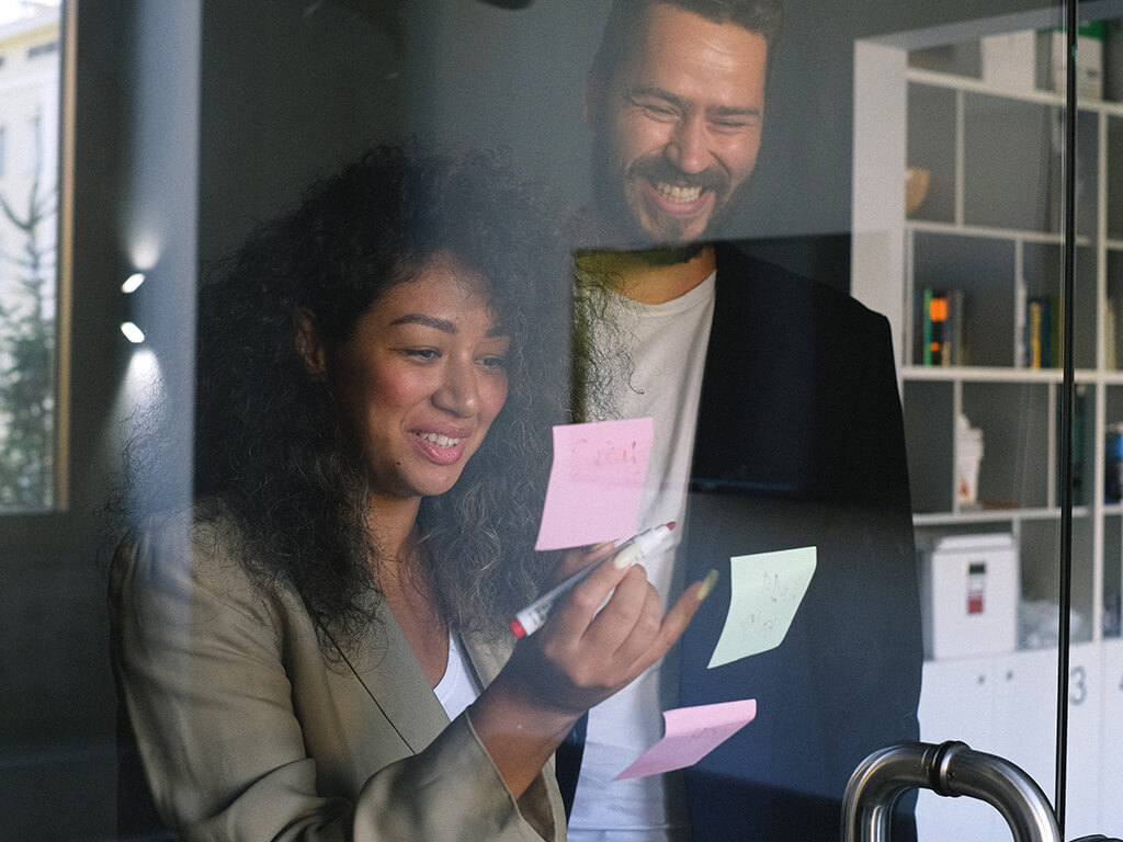 Desenvolvimento profissional para impulsionar a carreira e aumentar a renda. Homem e mulher sorrindo felizes, atuando como uma equipe, em ambiente corporativo, interagindo próximo a uma janela. A Mulher usa canetão para escrever em post-its fixados na porta de vidro.