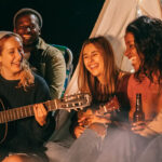 Equilibrando Diversão e Finanças: Estratégias para Momentos com Amigos dentro do Orçamento. Amigos felizes, tocando violão juntos na praia, durante a noite, desfrutando de bebidas e música.
